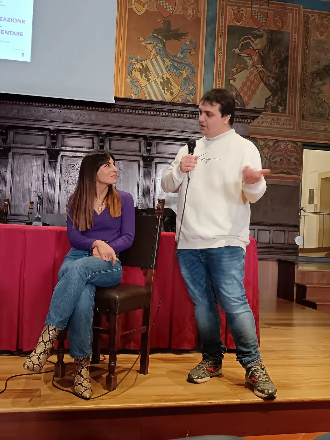 Francesco parla al microfono nella sala affrescata del Palazzo dei Notari di Perugia; Giorgia Bellini, seduta, lo ascolta
