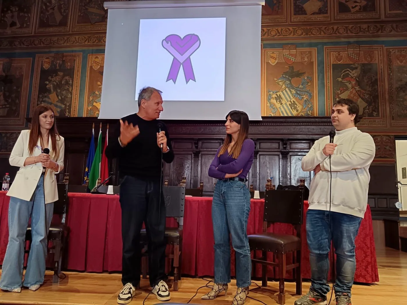Il palco della presentazione di Corabea al Palazzo dei Notari: quattro relatori in piedi con i microfoni sotto il logo proiettato; Francesco, a destra, in felpa bianca